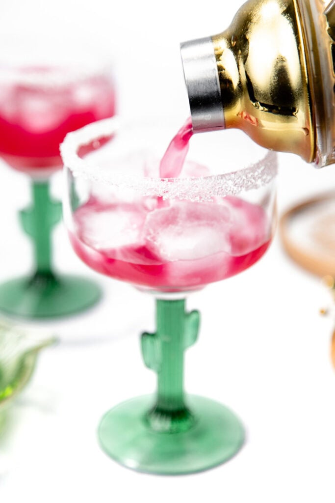 A gold shaker pours pink cocktail into a cactus-stem glass with a sugared rim and ice cubes.