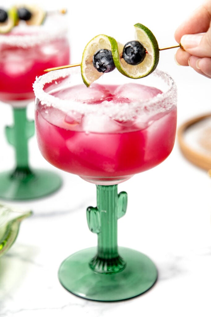 Pink margarita with ice in a cactus glass, garnished with lime slices and blueberries on a skewer.