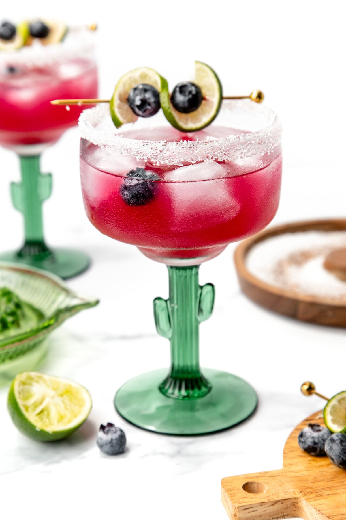 A purple cocktail with blueberries and lime in a cactus glass, garnished with salt and lime slices.