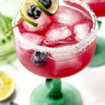 A pink cocktail with ice and blueberries, garnished with lime, in a glass with a salted rim.
