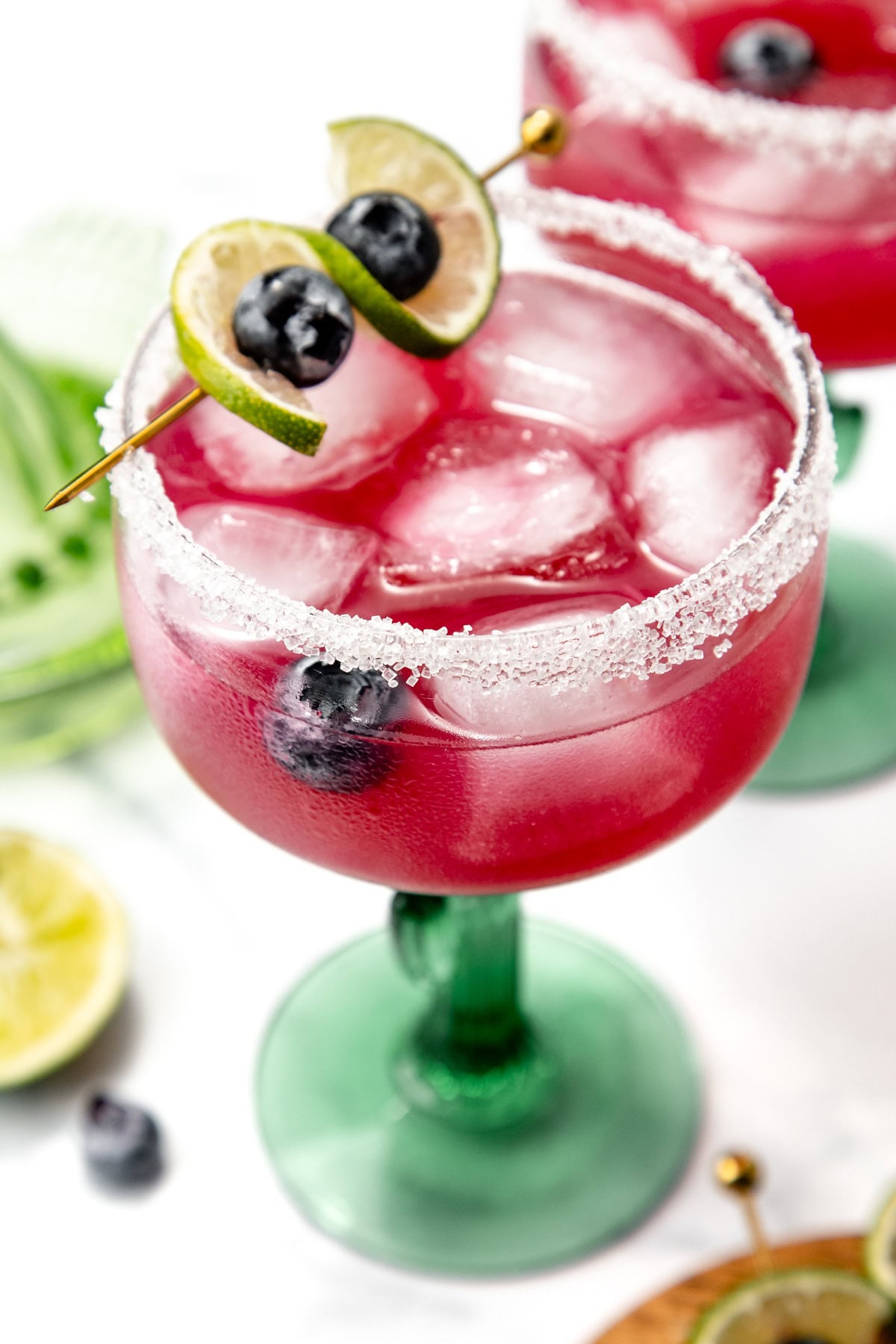 A pink cocktail with ice and blueberries, garnished with lime, in a glass with a salted rim.