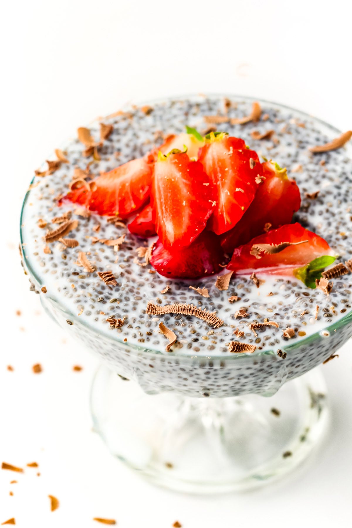 Chia pudding in a glass dish topped with sliced strawberries and chocolate shavings on white background.