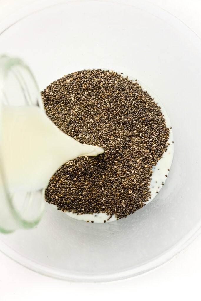 Milk being poured from a glass bottle over a pile of chia seeds in a clear mixing bowl.