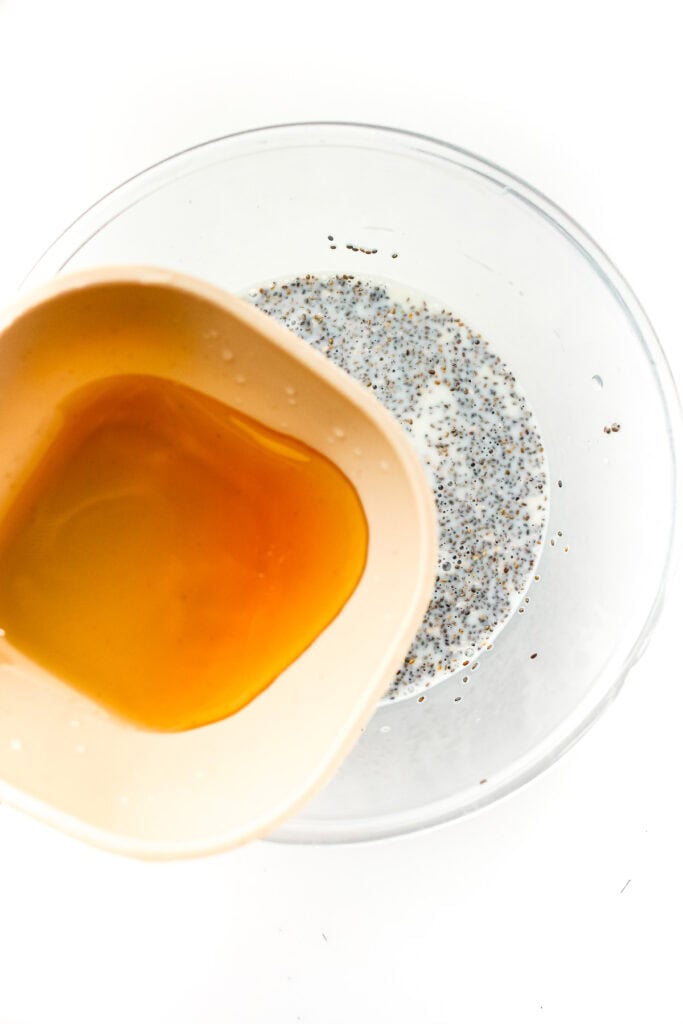 Honey being poured from a white bowl into a clear bowl with milk and poppy seeds, against a white background.