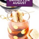 A cocktail in a polka dot glass with two sandwich squares on a skewer, labeled “Drinks for August.”.