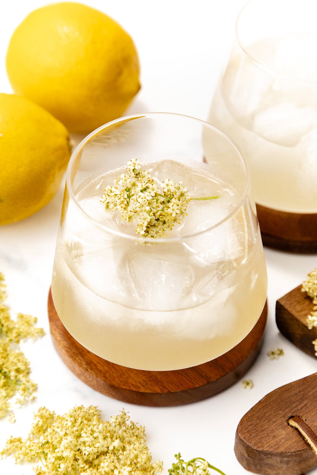 A glass of iced lemon drink garnished with elderflowers, with lemons and flowers nearby on a white surface.