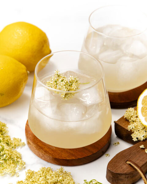 Two glasses of iced elderflower lemonade with lemons and elderflowers on a white surface.