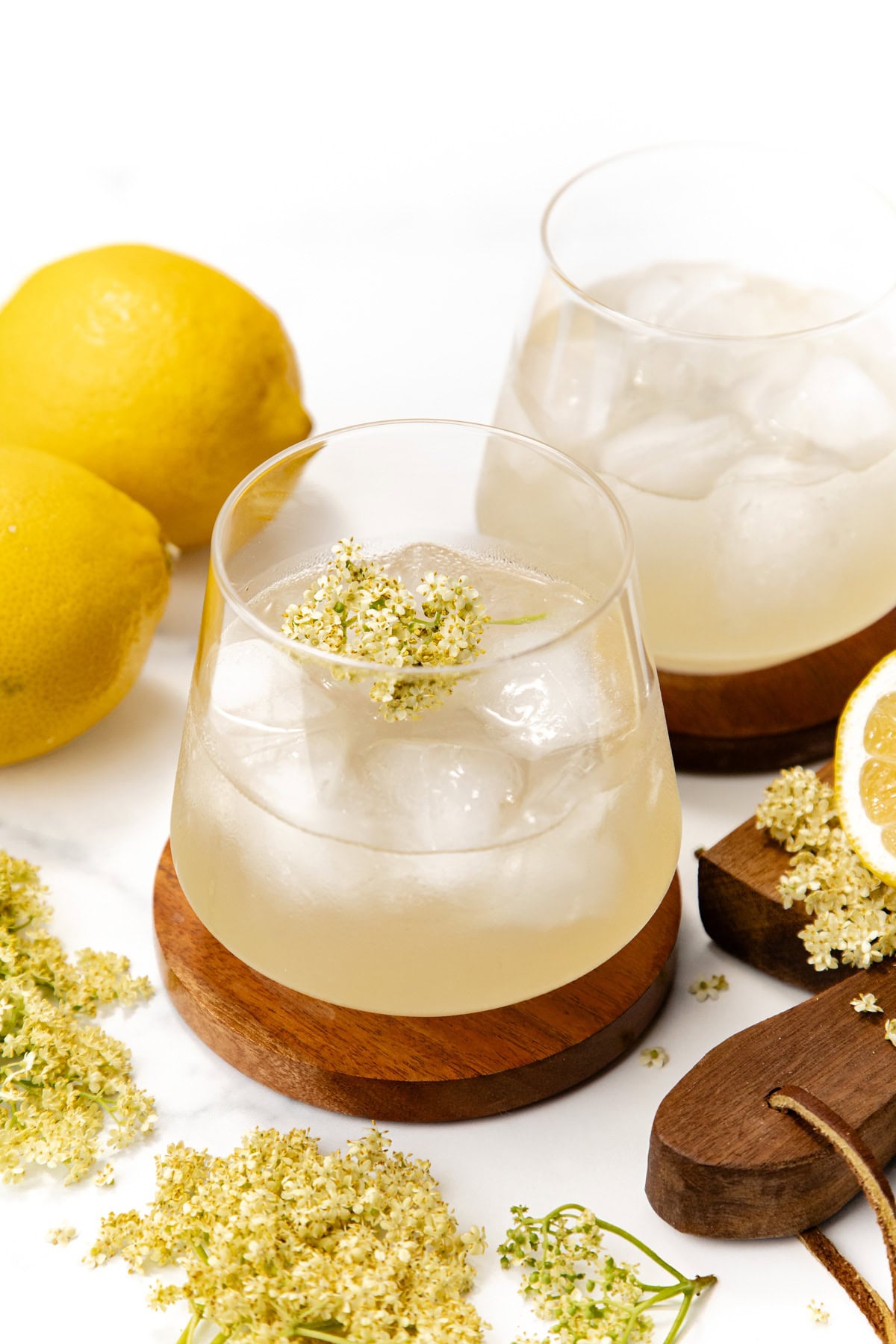 Two glasses of iced elderflower lemonade with lemons and elderflowers on a white surface.