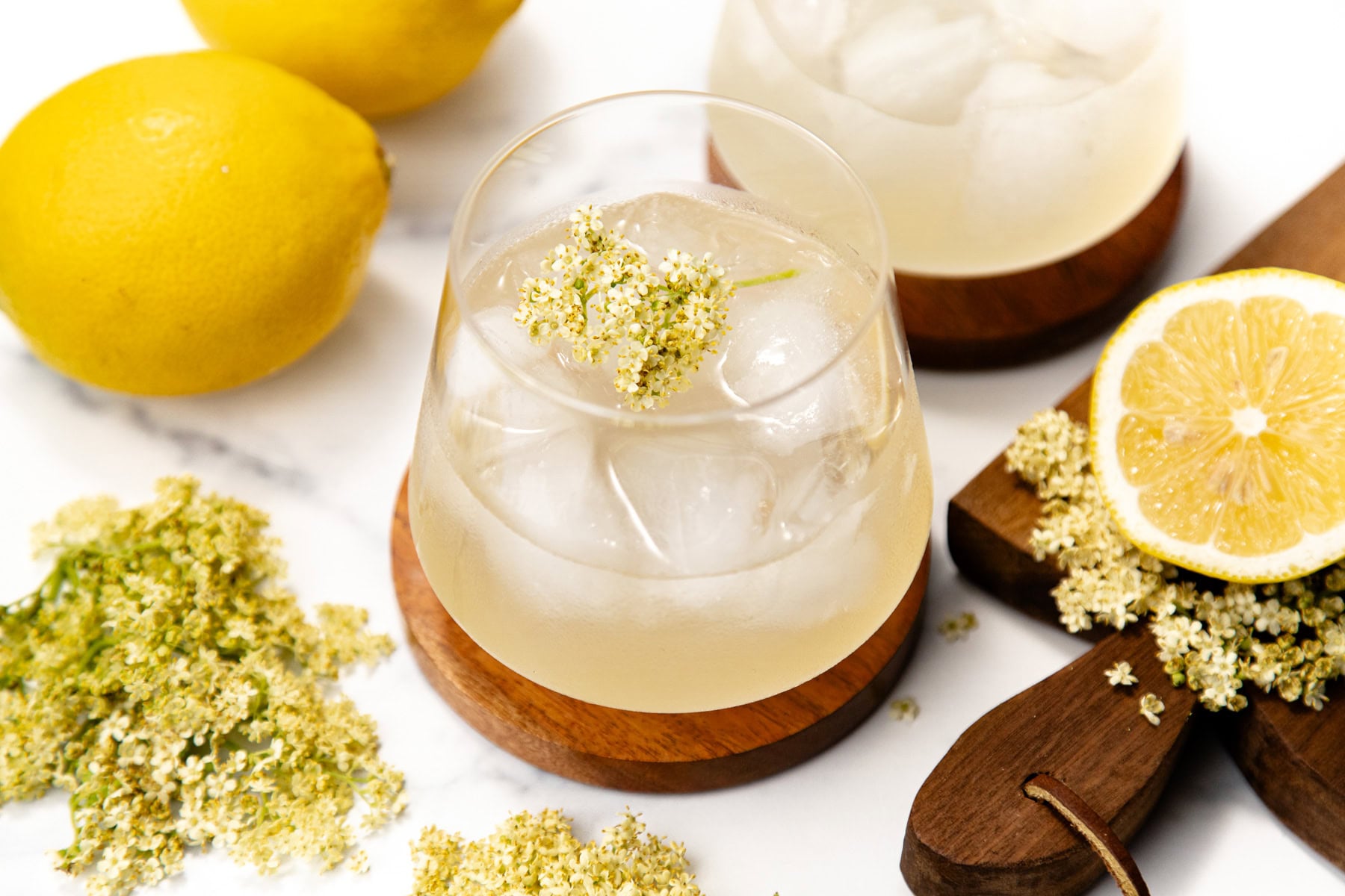 A glass of iced lemonade garnished with elderflowers, surrounded by fresh lemons and elderflowers.
