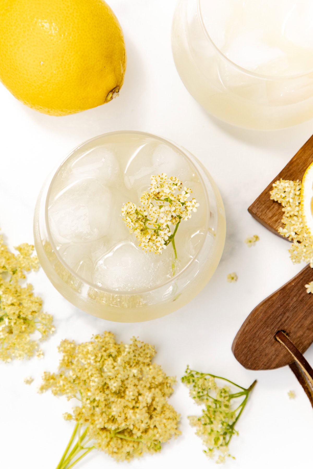 A glass of iced lemonade with elderflower, a lemon, elderflowers, and a wooden spoon on a white surface.