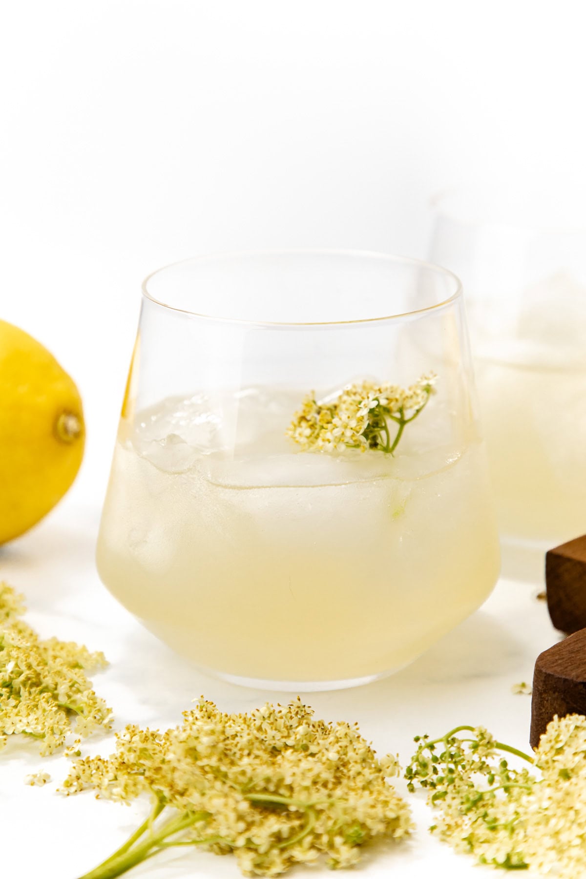 A glass of iced lemonade garnished with elderflower, with fresh elderflowers and a lemon nearby.