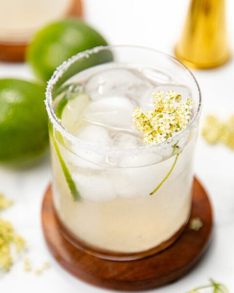 A cocktail with ice, lime slice, salted rim, and a small white flower, on a wooden coaster with limes nearby.