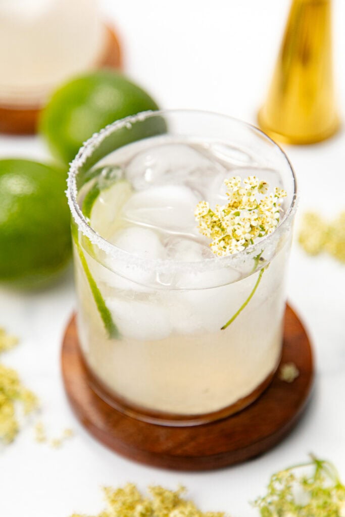 A cocktail with ice, lime slice, salted rim, and a small white flower, on a wooden coaster with limes nearby.