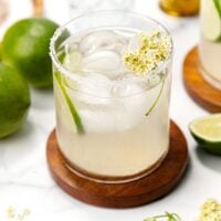A cocktail with ice, lime wedge, salt rim, and a flower garnish sits on a wooden coaster near fresh limes.