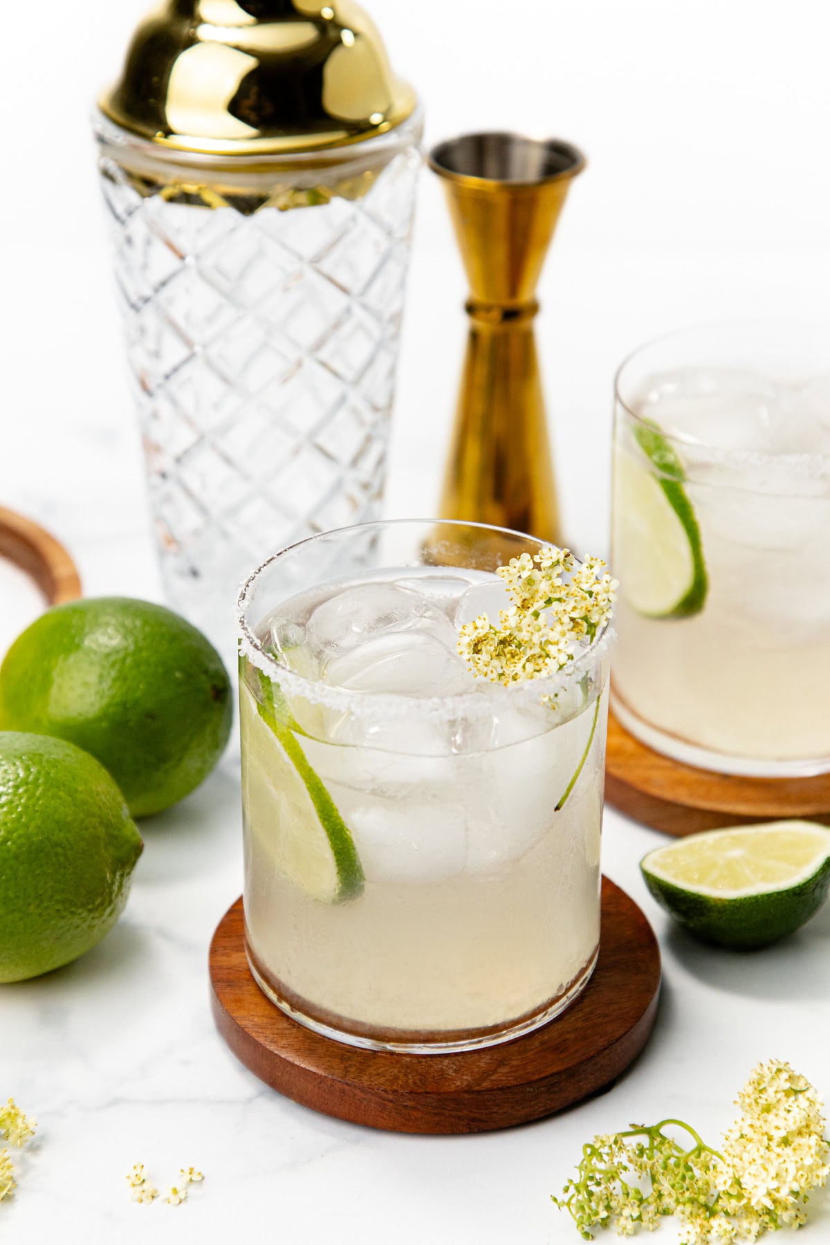 Two cocktails with ice, lime wedges, and flowers on coasters; shaker and jigger in background, limes nearby.