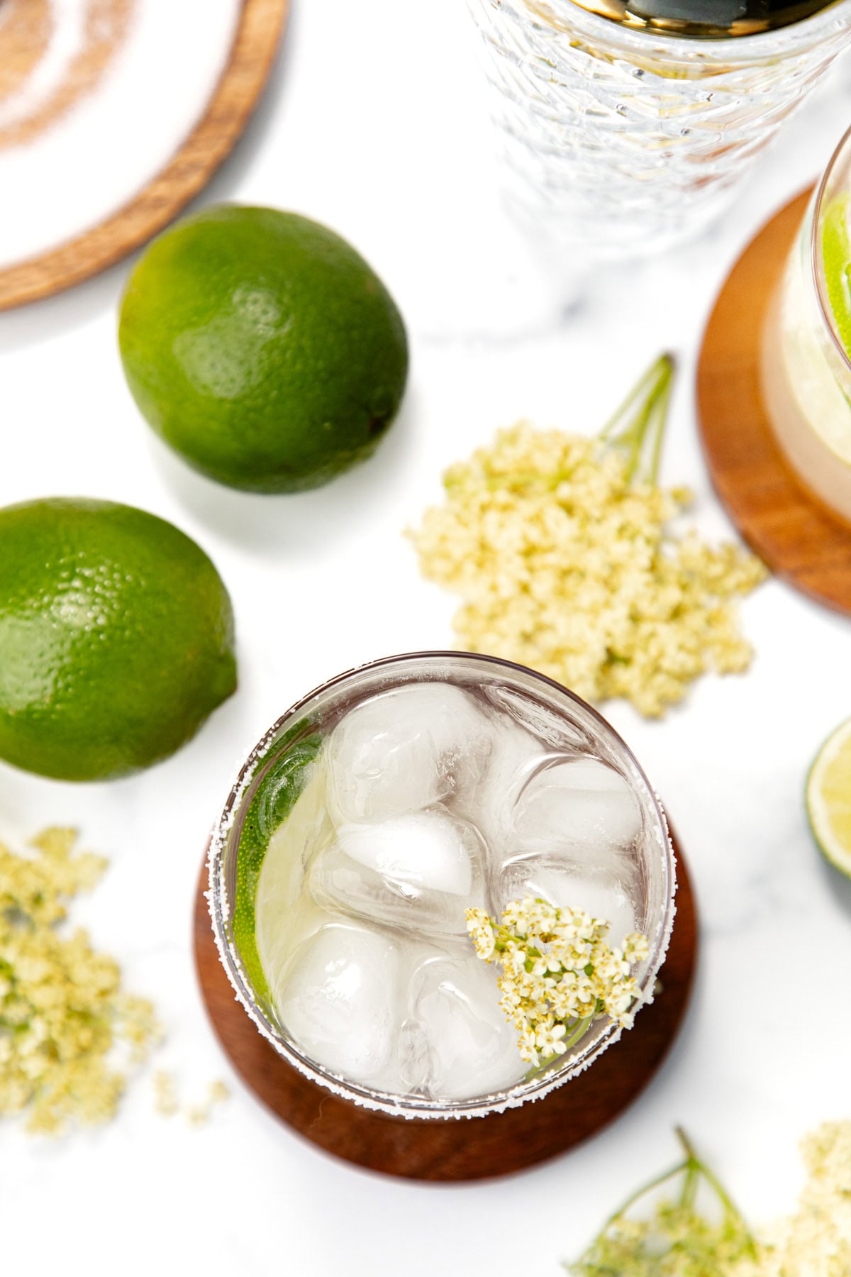 A glass of iced drink garnished with elderflowers, surrounded by limes and elderflowers on a white surface.