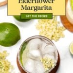 A glass of elderflower margarita with ice, lime, and elderflowers, next to a recipe card.