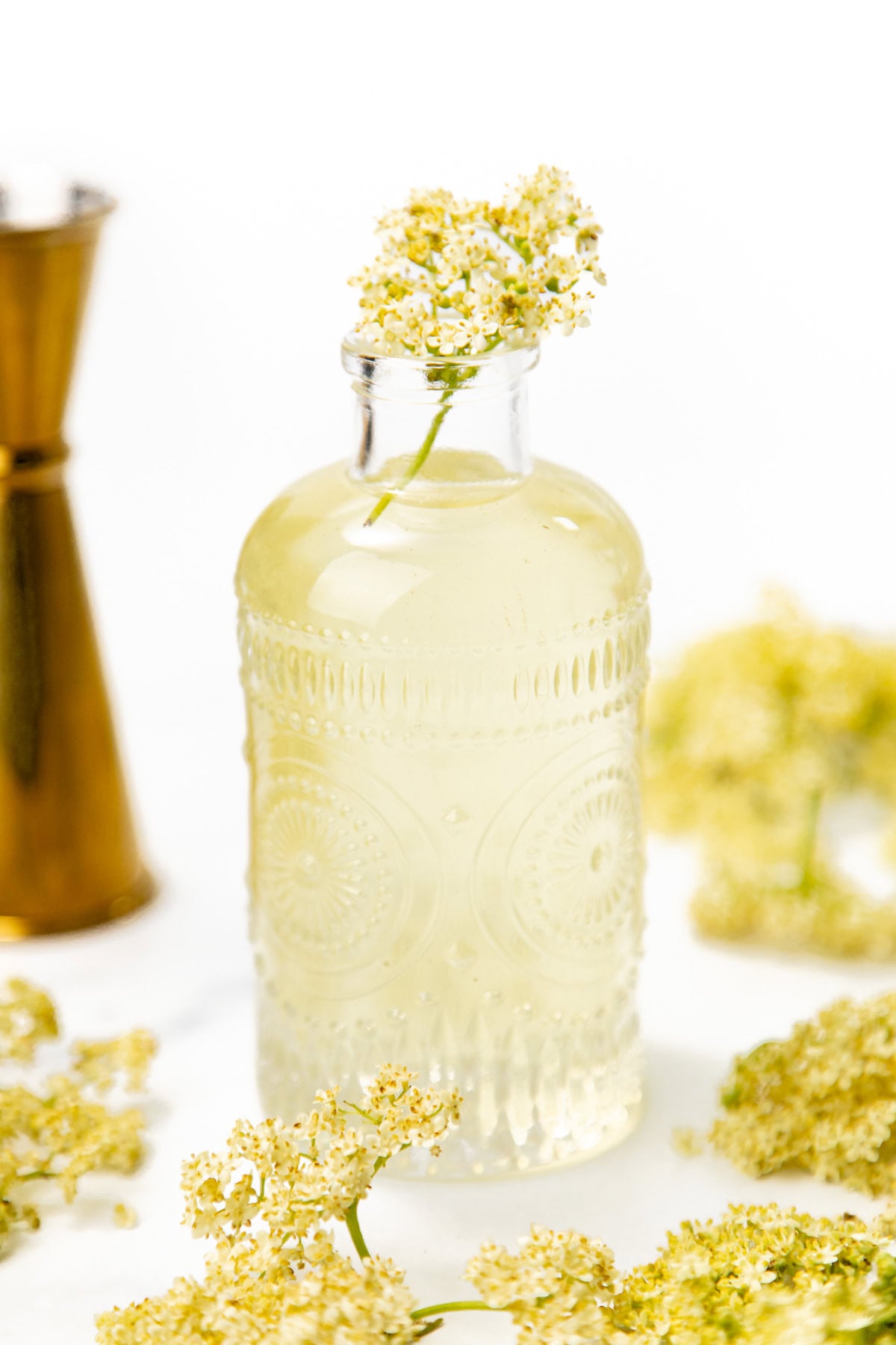A glass bottle of elderflower cordial with a sprig of elderflower, surrounded by loose blossoms.