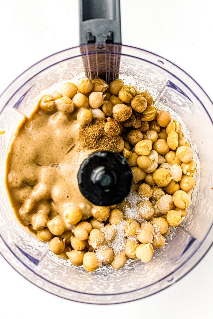 Overhead view of chickpeas, tahini, spices, and salt in a food processor ready to be blended.