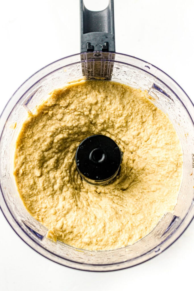 A food processor filled with creamy, blended hummus on a white background.