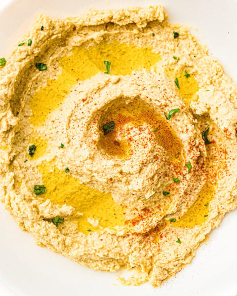 Creamy hummus spread on a plate, drizzled with olive oil, sprinkled with paprika and chopped parsley.