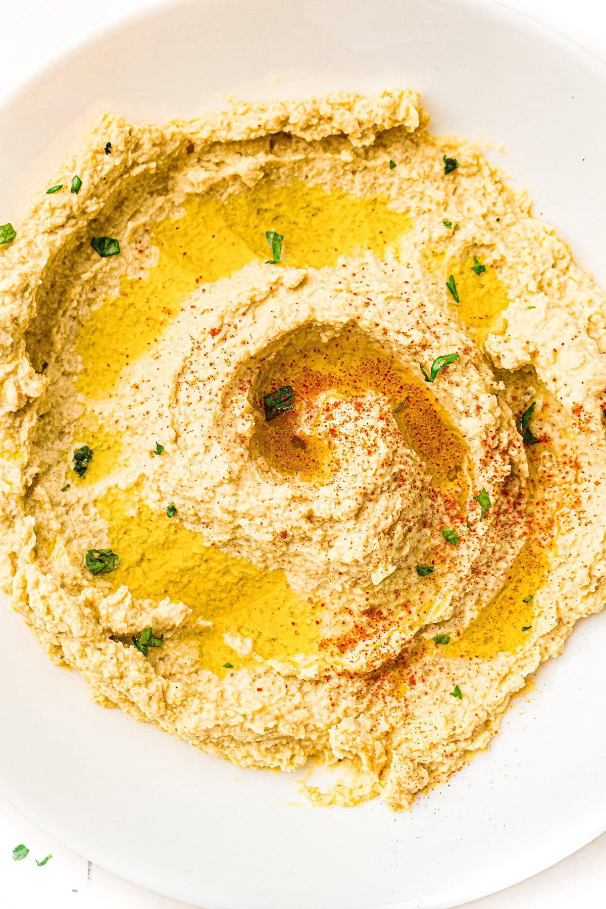 Creamy hummus spread on a plate, drizzled with olive oil, sprinkled with paprika and chopped parsley.