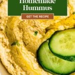 Creamy homemade hummus garnished with olive oil, herbs, and cucumber slices in a white bowl.