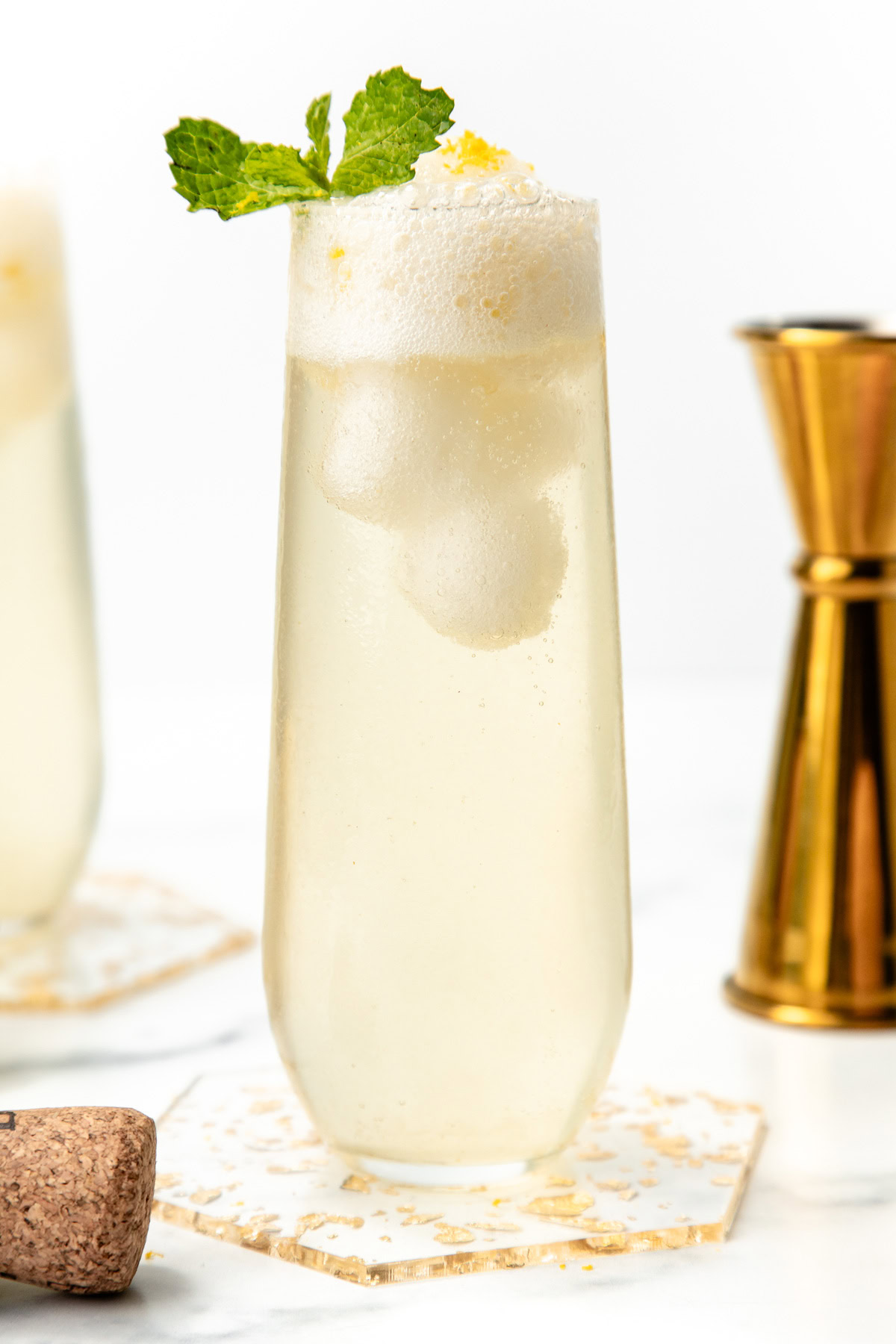 Tall glass of fizzy lemonade with ice, mint garnish, and lemon zest on top, next to a gold jigger.