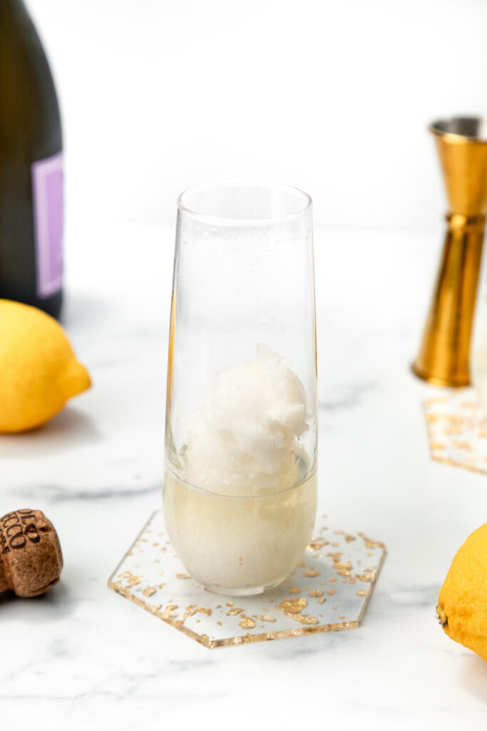 A clear glass with a scoop of sorbet, surrounded by lemons, champagne, and bar tools on a marble surface.