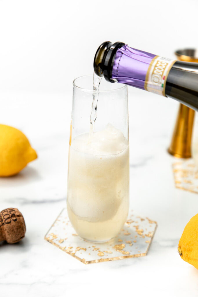 A drink is being poured into a glass with ice, surrounded by lemons and a cork on a white surface.