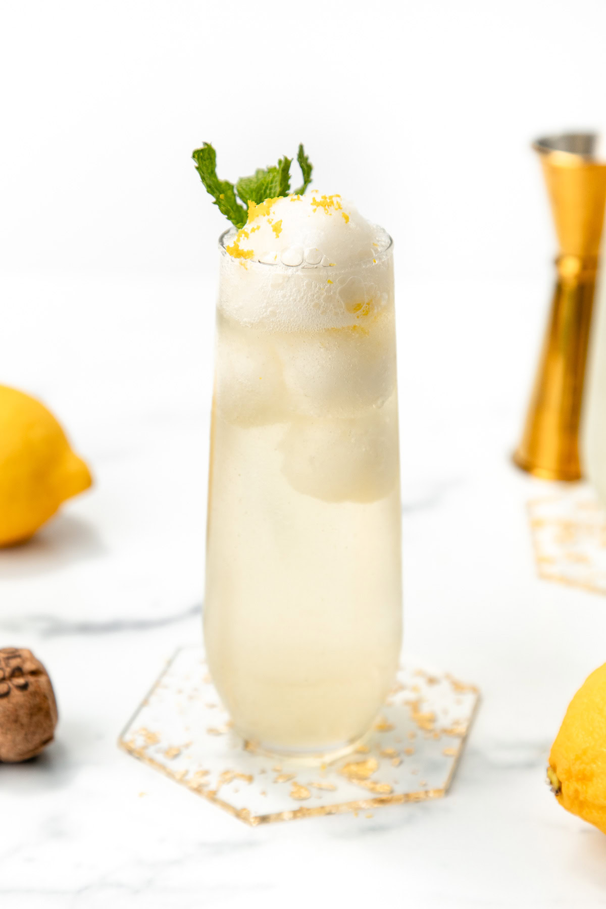 Tall glass of fizzy lemonade with ice, foam on top, lemon zest, and a mint sprig garnish on a marble surface.