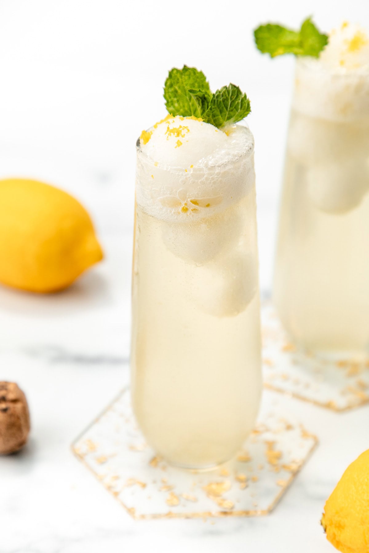 A tall glass of lemon fizz drink with ice cream, garnished with mint and lemon zest, on a marble surface.