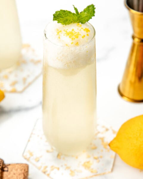 A tall glass with a frothy lemon drink, garnished with mint and lemon zest, on a marble surface.