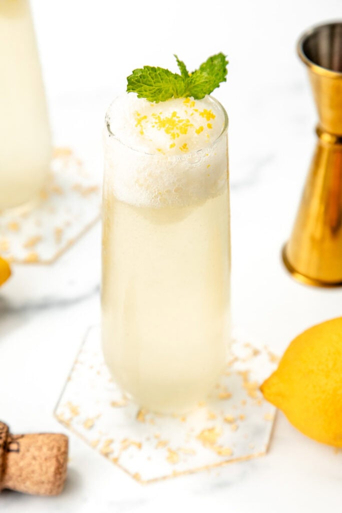 A tall glass with a frothy lemon drink, garnished with mint and lemon zest, on a marble surface.
