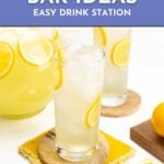 Two glasses of lemonade with ice and lemon slices on coasters, next to a pitcher and a lemon.