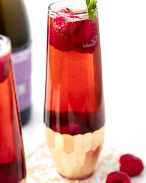 A tall glass of red sparkling drink with raspberries and mint, next to loose raspberries and a purple bottle.