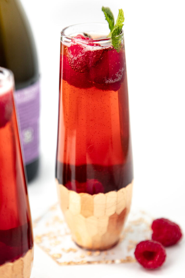 A tall glass of red sparkling drink with raspberries and mint, next to loose raspberries and a purple bottle.