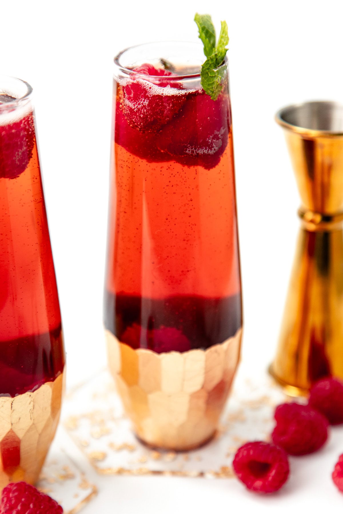A tall glass of red sparkling drink with raspberries, gold base, and mint garnish, next to fresh raspberries.