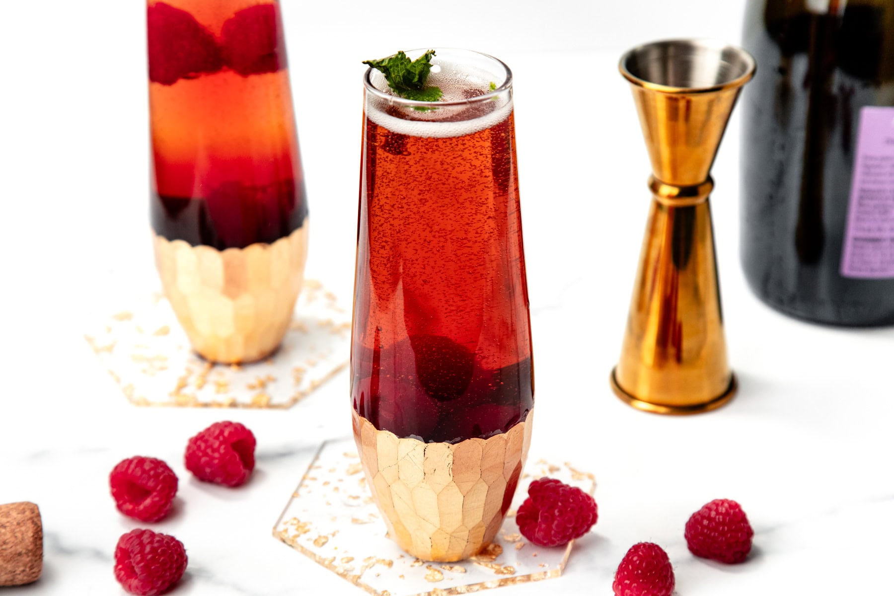 A tall glass of sparkling red drink with raspberries and mint, next to gold barware and fresh raspberries.