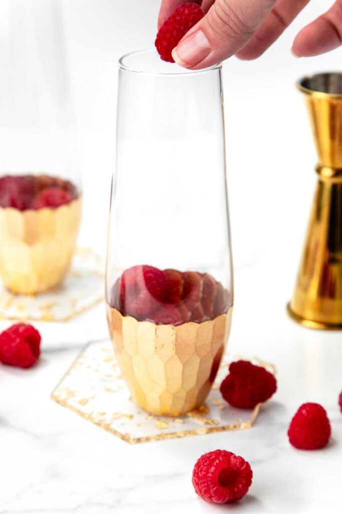 A hand drops a raspberry into a gold-accented glass with more raspberries inside, on a marble surface.