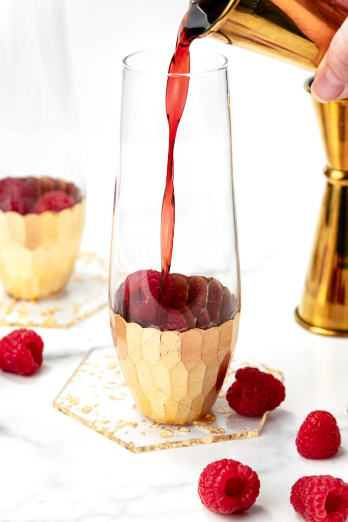 Red liquid being poured into a gold-accented glass with raspberries on a marble surface.