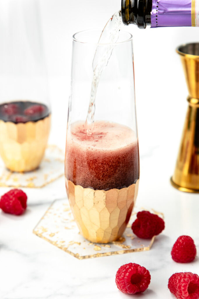 Champagne is being poured into a flute with raspberries inside, surrounded by loose raspberries.