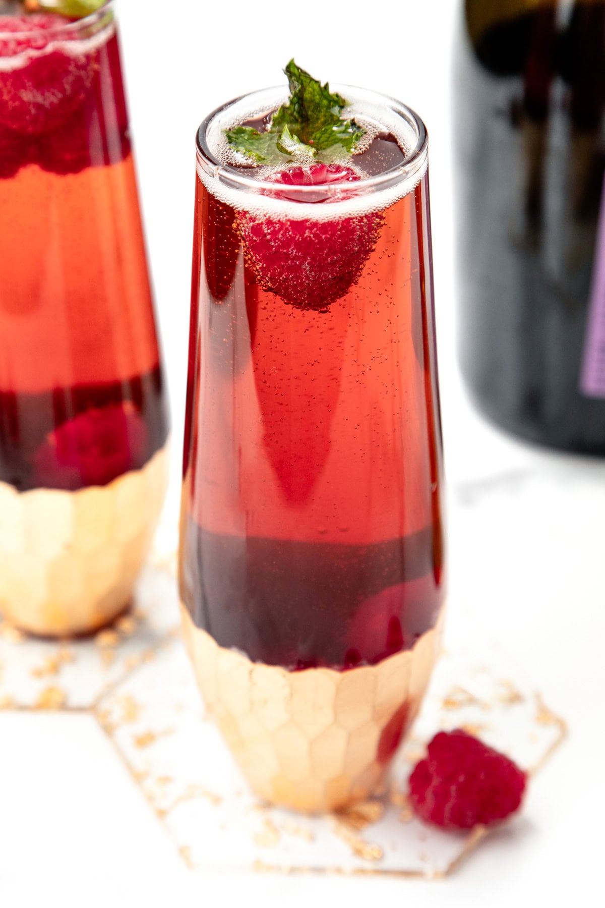 Tall glass of red sparkling drink garnished with a raspberry and mint, with a bottle and coaster nearby.