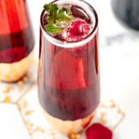 A glass of red sparkling drink with raspberries and a mint garnish on a white surface.
