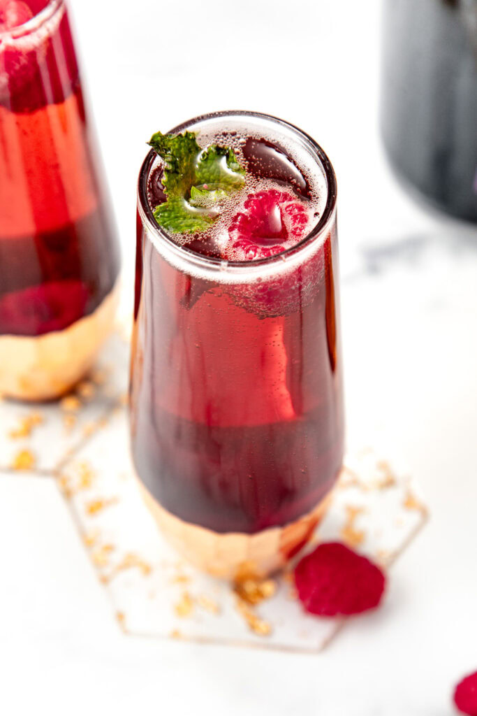 A glass of red sparkling drink with raspberries and a mint garnish on a white surface.