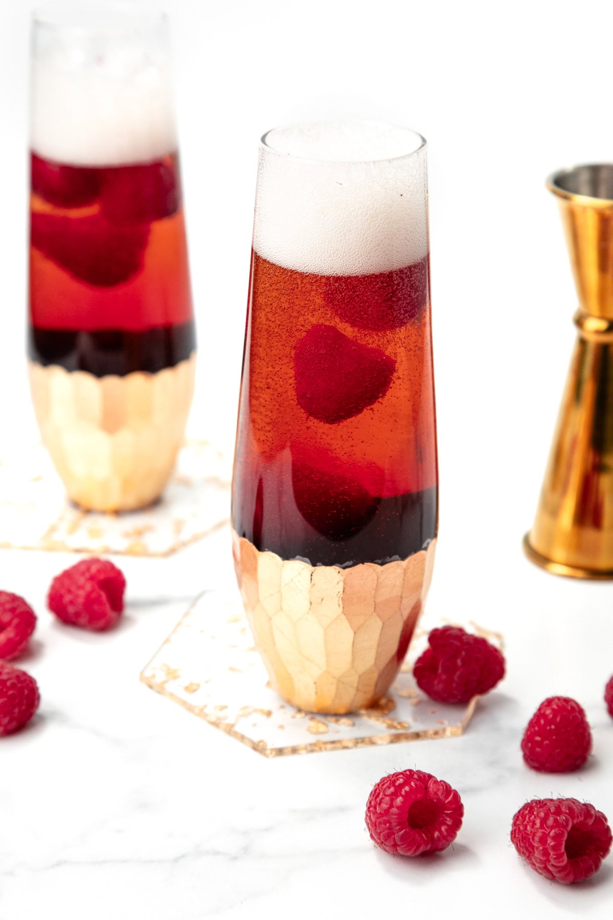Two champagne flutes with red sparkling drink, foam on top, and raspberries, surrounded by fresh raspberries.