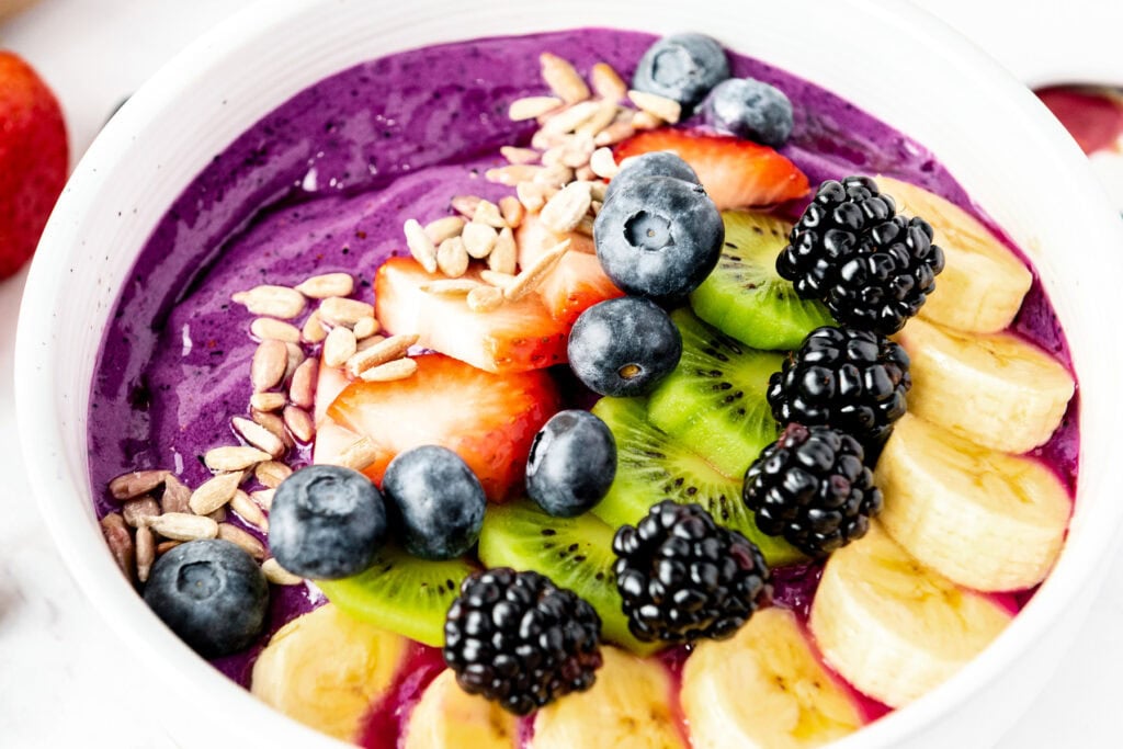 A smoothie bowl topped with banana, kiwi, strawberries, blackberries, blueberries, and sunflower seeds.