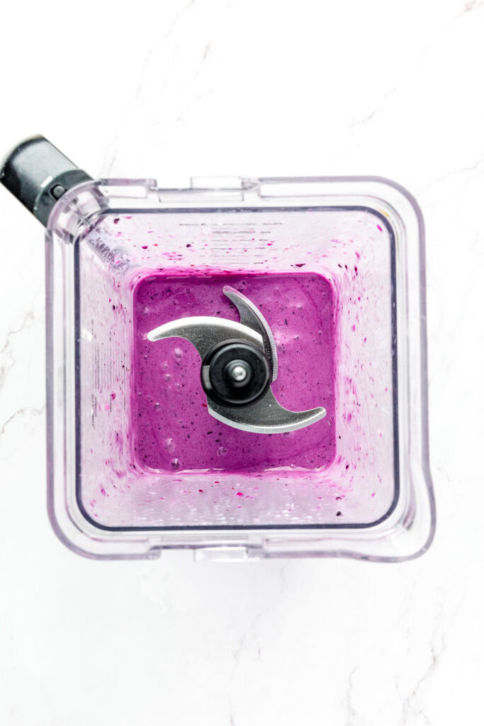 Top view of a blender with bright purple smoothie mixture inside on a white marble surface.