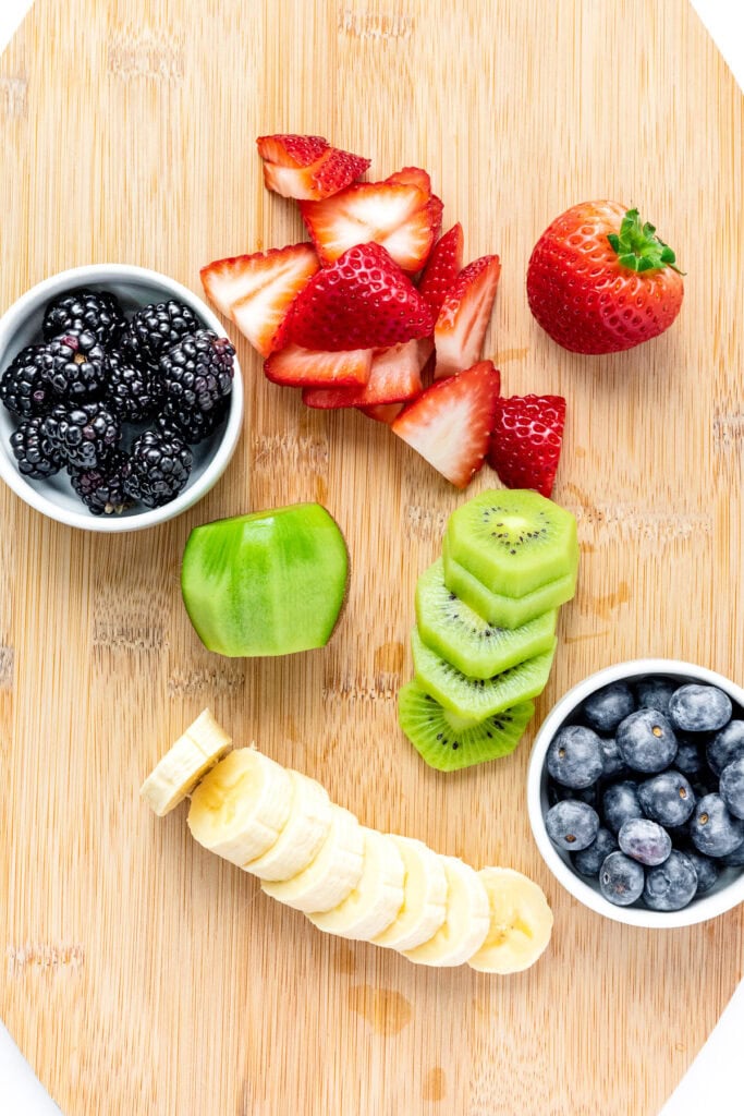 Sliced strawberries, blackberries, kiwi, banana, and blueberries on a wooden cutting board.
