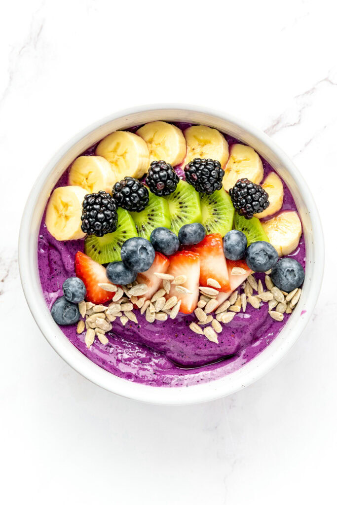 A smoothie bowl topped with banana, kiwi, blackberries, blueberries, strawberries, and sunflower seeds.
